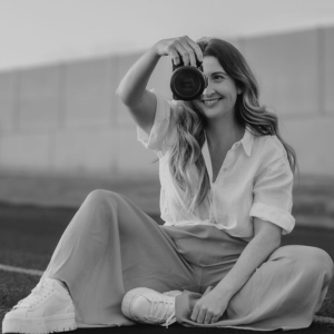 A black and white photo of Emily holding a camera while sitting crosslegged on the road