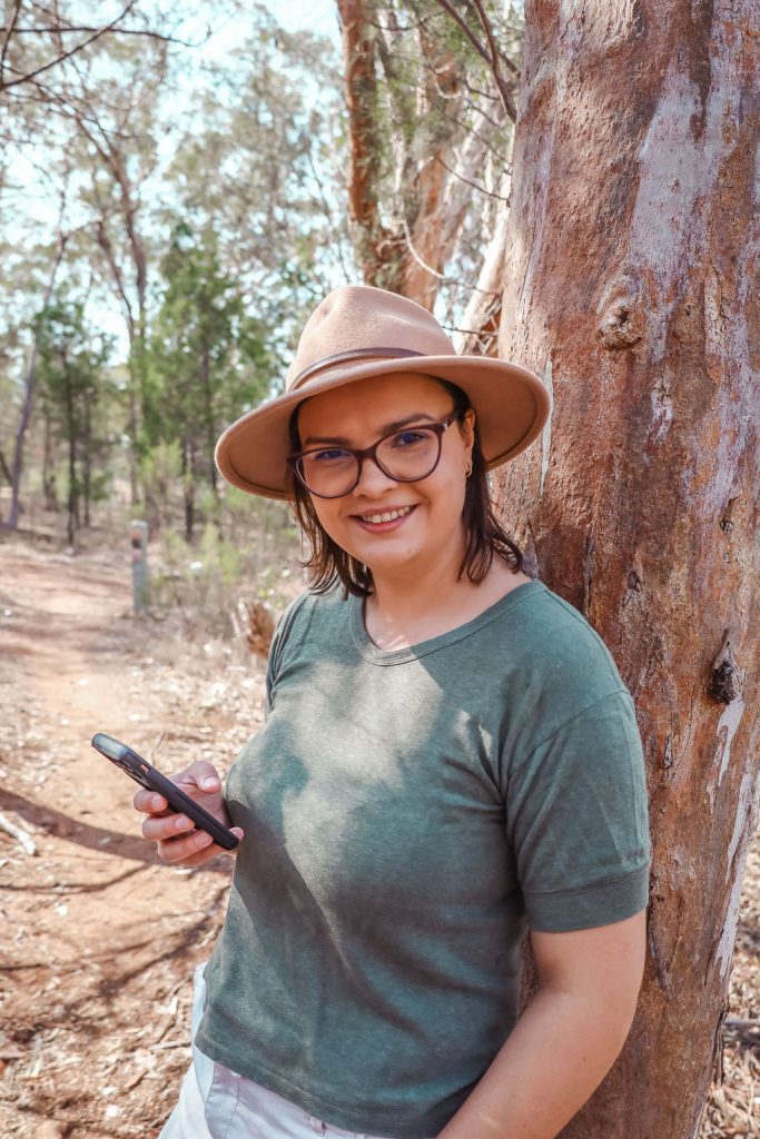 A woman wearing a hat and glasses standing next to a tree, holding a phone smiling and looking directly into the camera.