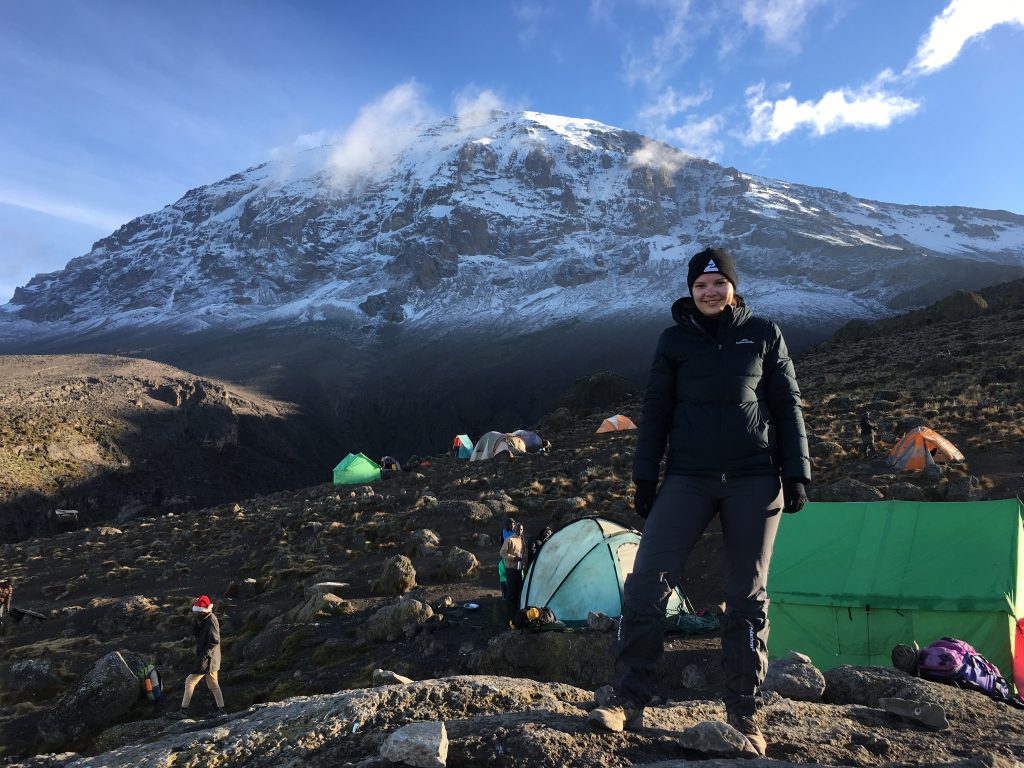 A hiker posing in front of a snow-capped mountain with a base camp in the foreground.