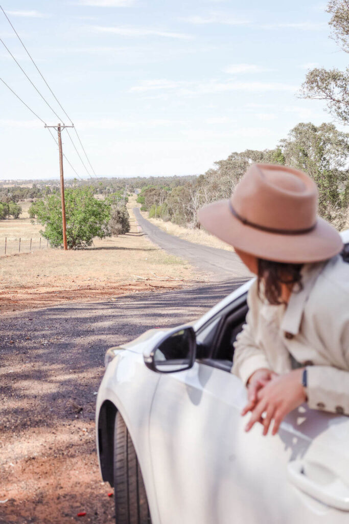 Person in a hat leans out of a car window, looking at a rural road stretching into the distance, surrounded by fields and trees.