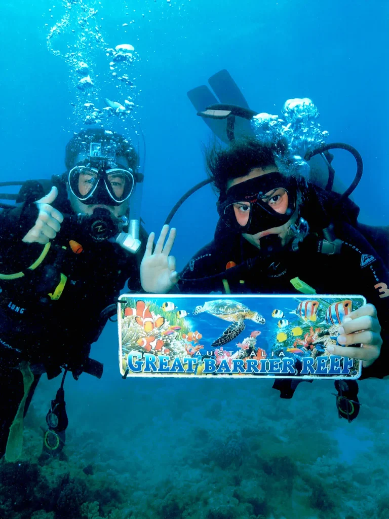 Two scuba divers underwater holding a "Great Barrier Reef" sign, surrounded by clear blue water and coral.